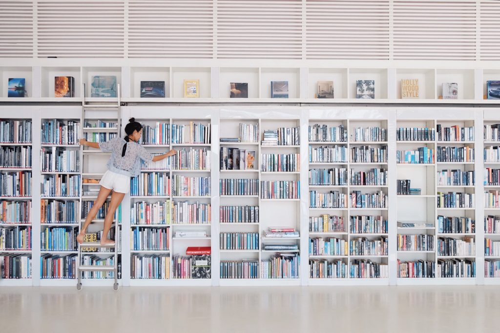 Woman on a ladder reaching for a book on a bookshelf.