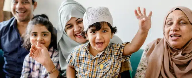 A family sitting on a couch together waving and smiling at the camera.