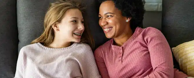A white preteen girl and black woman smile at each other on a couch