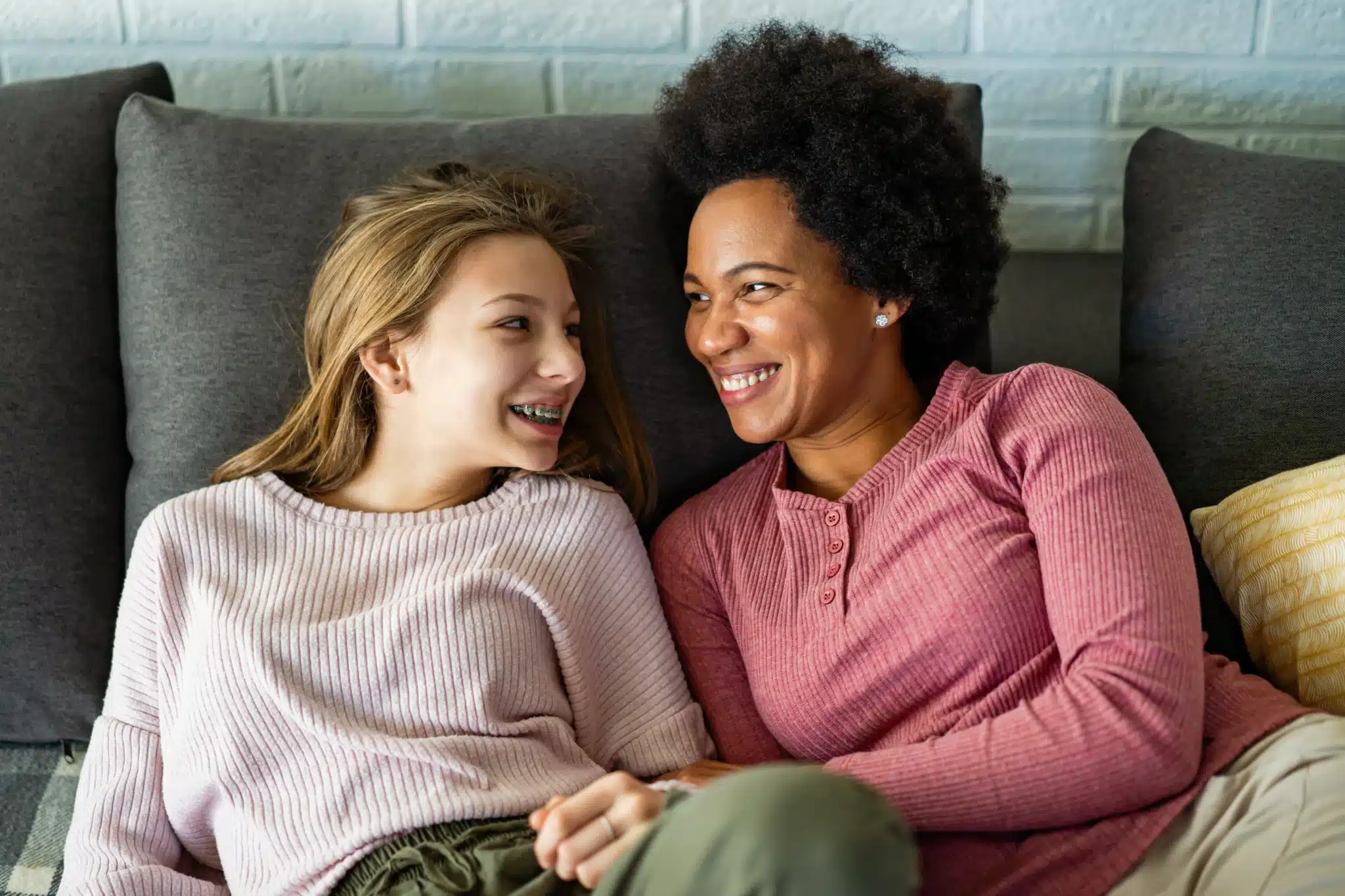 A white preteen girl and black woman smile at each other on a couch