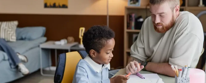 Side view of little boy drawing with colorful felt tip pens while supportive dad helps his son sitting at desk together in kids room