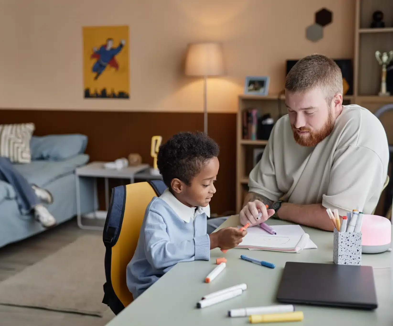 Side view of little boy drawing with colorful felt tip pens while supportive dad helps his son sitting at desk together in kids room