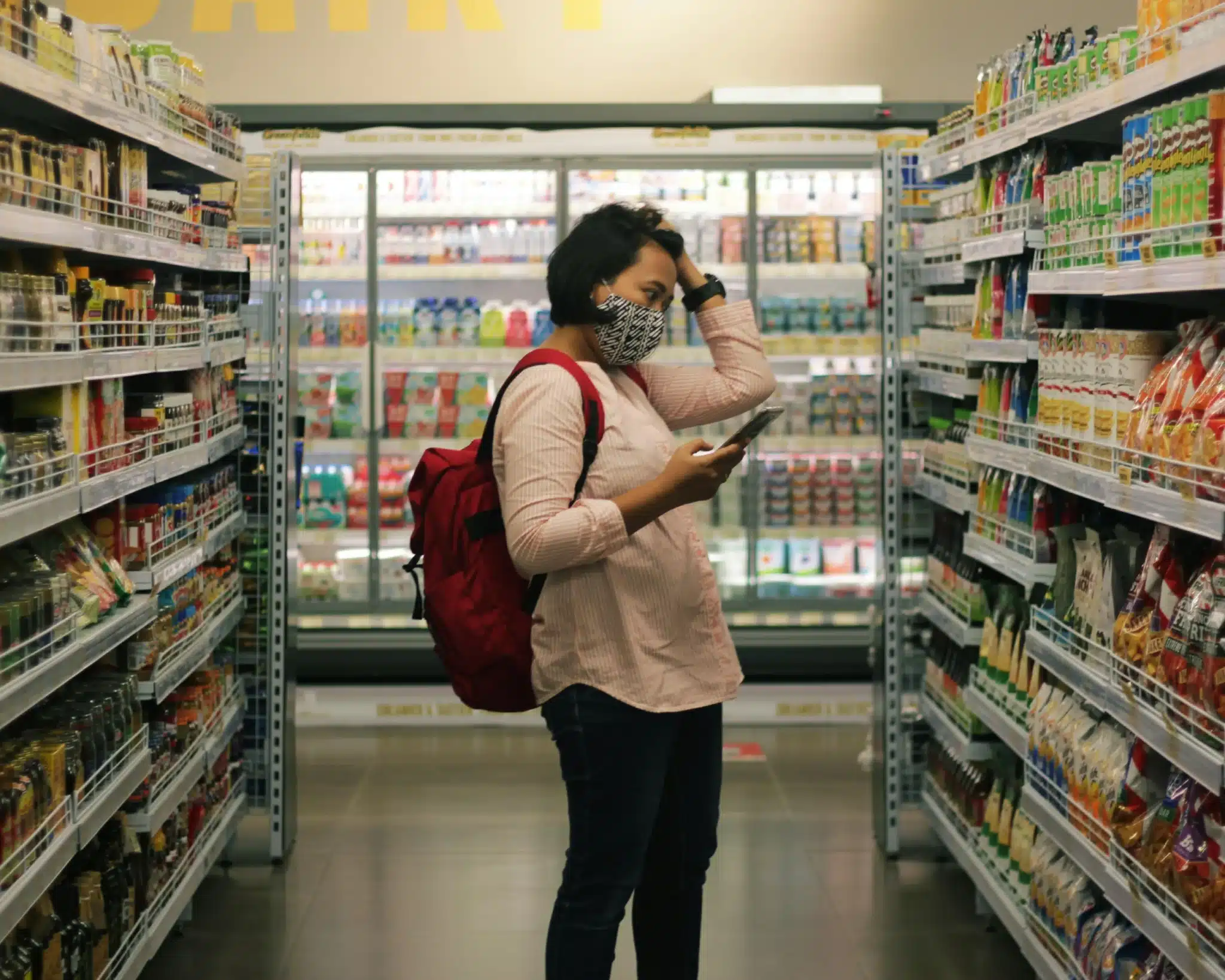 A woman wearing a face mask and a backpack is standing in a grocery store aisle examining a product with her left hand on top of her head
